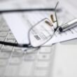 Brille und Stift auf ausgefülltem Steuerformular, daneben ein Laptop, Symbol für digitale Steuerbearbeitung