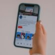 Hand hält ein Smartphone, auf dessen Bildschirm ein Social Media Feed dargestellt wird, Fokus auf moderner digitaler Kommunikation