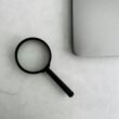 Eine Lupe liegt neben einem aufgeklappten Laptop auf einem Tisch, ideal für Themen rund um Recherche oder SEO-Analyse.