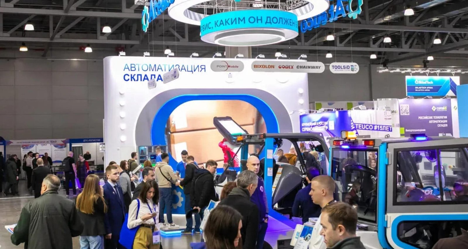 Besucher schlendern auf einer Ausstellung durch eine Messehalle mit zahlreichen Ständen und digitalen Displays.