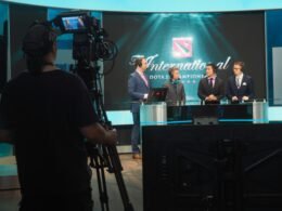 Drei Männer im Anzug stehen bei einer Dota 2 Competition vor Kameras und werden gefilmt