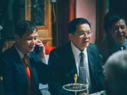 Drei Männer in Anzügen stehen bei einer formellen Zusammenkunft und unterhalten sich in geschäftlicher Atmosphäre.