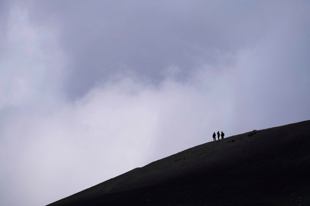 Drei Wanderer gehen einen dunklen, wolkenverhangenen Berghang hinauf, umgeben von dichter Natur und Felsen.