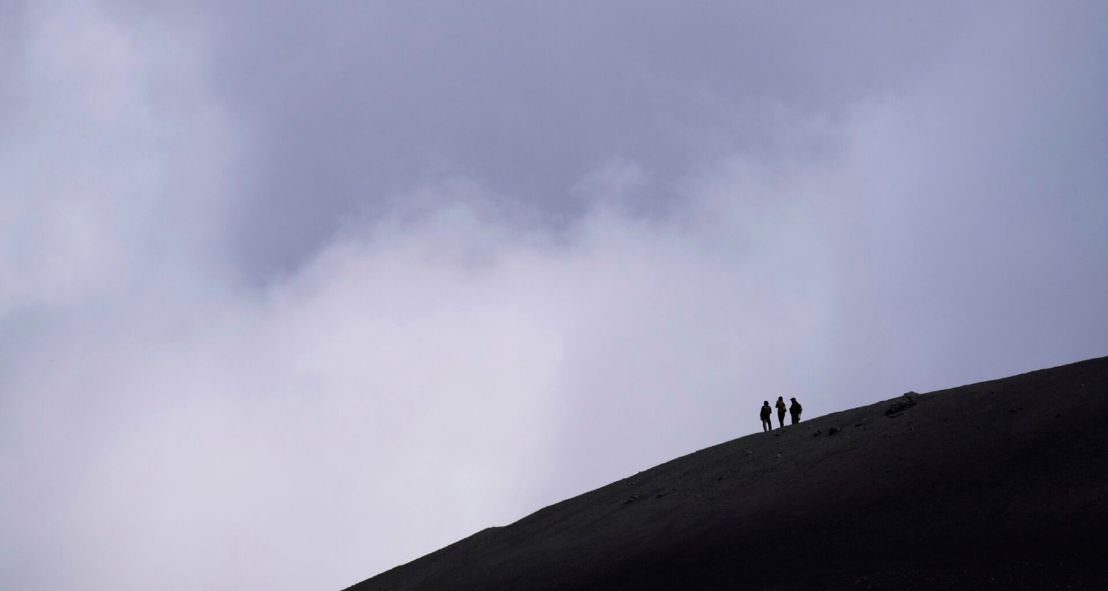 Drei Wanderer gehen einen dunklen, wolkenverhangenen Berghang hinauf, umgeben von dichter Natur und Felsen.