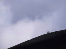 Drei Wanderer gehen einen dunklen, wolkenverhangenen Berghang hinauf, umgeben von dichter Natur und Felsen.