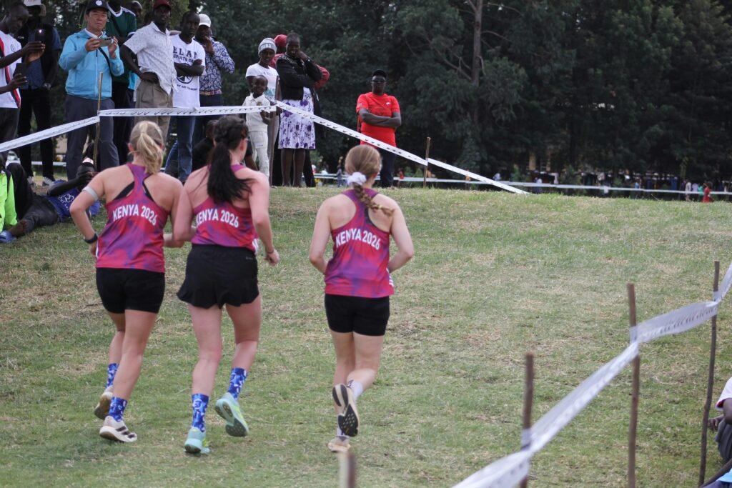 Drei Frauen in pinken Laufshirts rennen nebeneinander über ein grünes Grasfeld, sportlich aktiv im Freien.