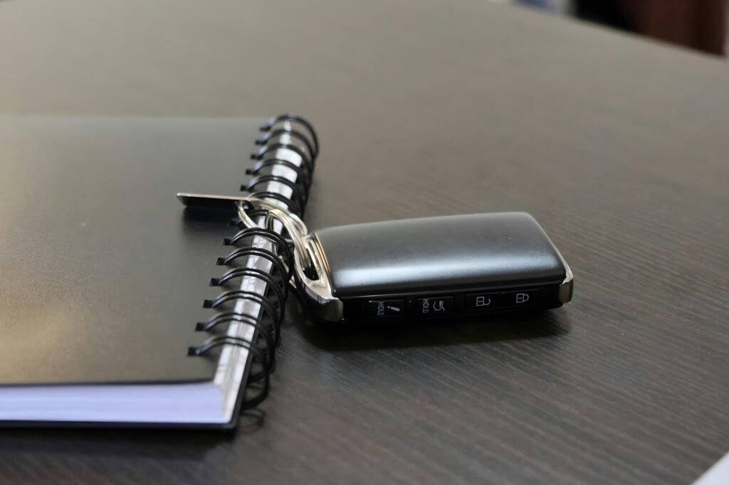 Ein Autoschlüssel und ein Notizbuch liegen auf einer hellen Oberfläche nebeneinander, deutliche Details beider Objekte sind erkennbar.