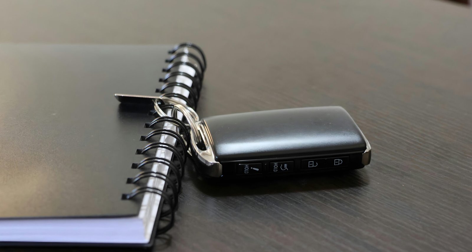 Ein Autoschlüssel und ein Notizbuch liegen auf einer hellen Oberfläche nebeneinander, deutliche Details beider Objekte sind erkennbar.