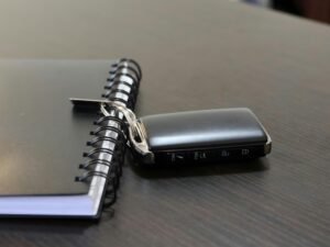 Ein Autoschlüssel und ein Notizbuch liegen auf einer hellen Oberfläche nebeneinander, deutliche Details beider Objekte sind erkennbar.