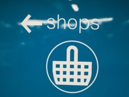 Blaues Schild mit weißem Einkaufskorb-Symbol als Zeichen für Onlineshopping, aufgenommen vor neutralem Hintergrund