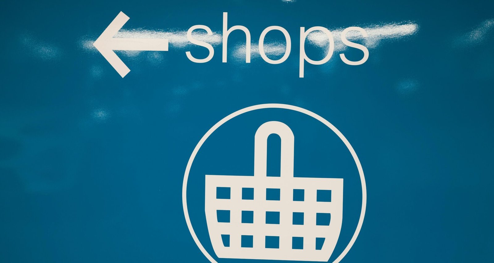 Blaues Schild mit weißem Einkaufskorb-Symbol vor schlichtem Hintergrund, als Piktogramm für Einkaufen oder Online-Shops geeignet.