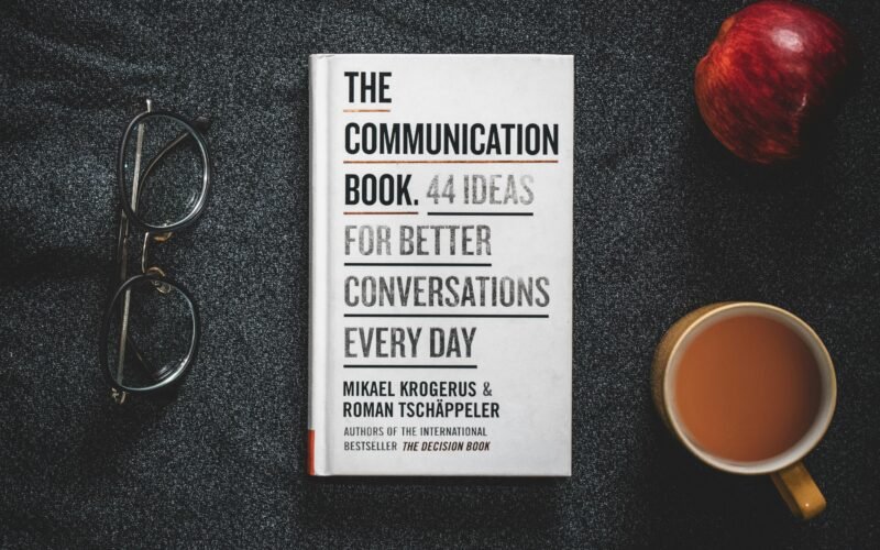 Ein aufgeschlagenes Buch und eine Tasse Kaffee stehen nebeneinander auf einem Holztisch in ruhiger Umgebung