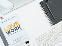 Ein Buch liegt auf einem Schreibtisch direkt neben einer Computertastatur, typischer Arbeitsplatz im Büroalltag.