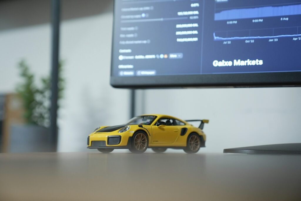 Gelbes Spielzeugauto steht auf einem Tisch vor einem eingeschalteten Computermonitor in einem modernen Arbeitsumfeld.