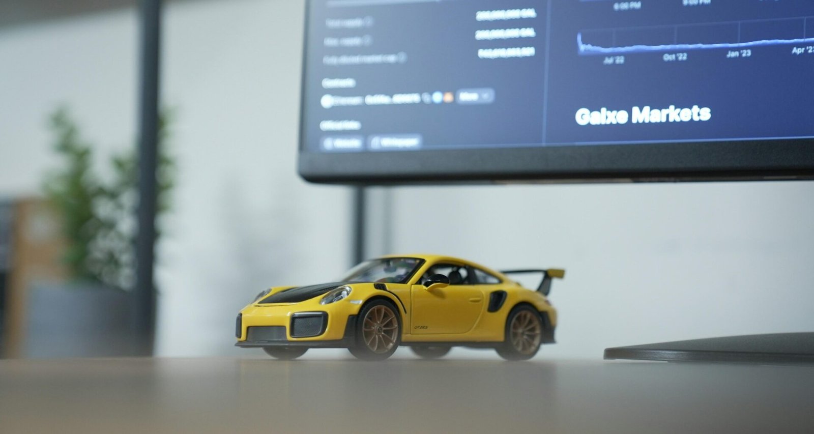 Gelbes Spielzeugauto steht auf einem Tisch vor einem eingeschalteten Computermonitor in einem modernen Arbeitsumfeld.