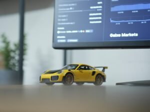Gelbes Spielzeugauto steht auf einem Tisch vor einem eingeschalteten Computermonitor in einem modernen Arbeitsumfeld.