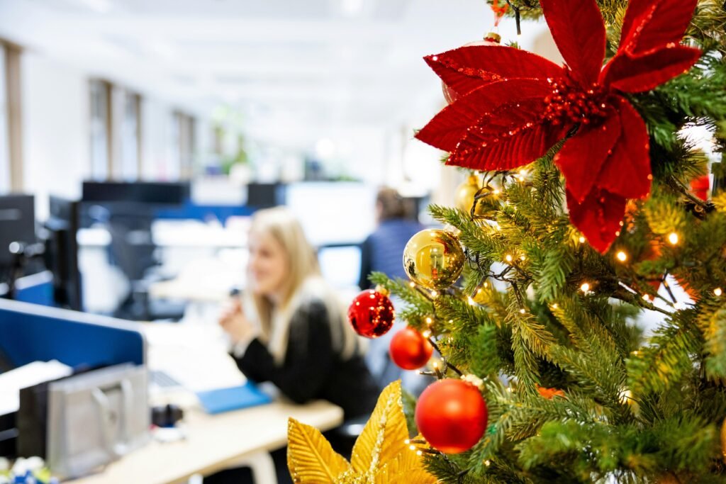 Ein geschmückter Weihnachtsbaum steht in einem Büro-Kubikel neben typischen Arbeitsmaterialien und bringt festliche Atmosphäre.