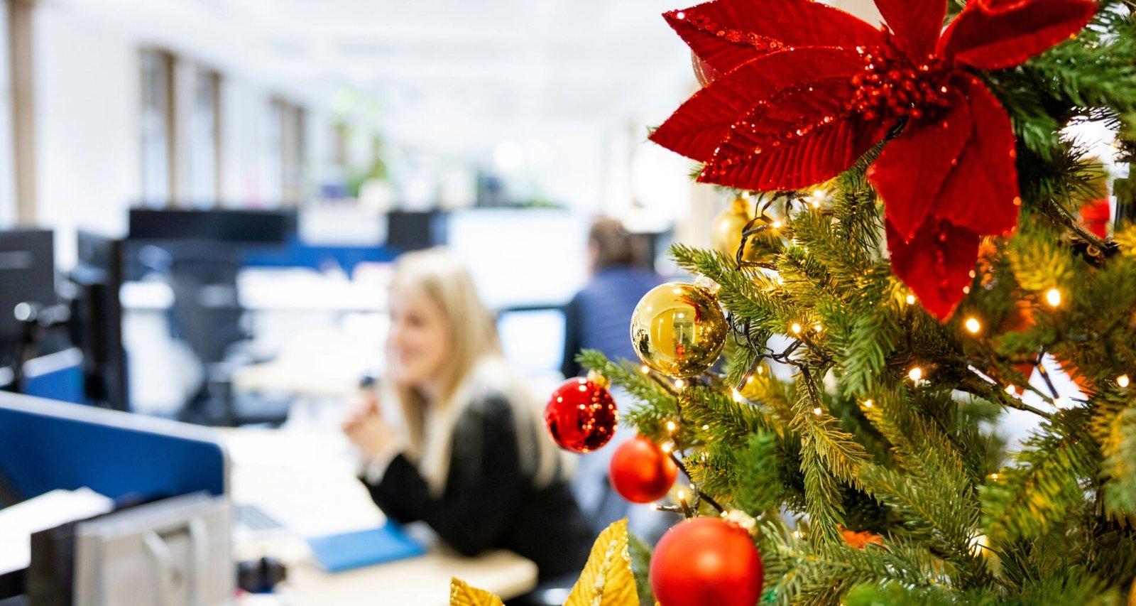 Ein geschmückter Weihnachtsbaum steht in einem Büro-Kubikel neben typischen Arbeitsmaterialien und bringt festliche Atmosphäre.