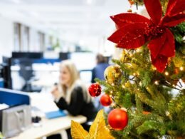 Ein geschmückter Weihnachtsbaum steht in einem Büro-Kubikel neben typischen Arbeitsmaterialien und bringt festliche Atmosphäre.