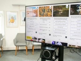 Großer Bildschirm an der Wand in einem modern eingerichteten Raum mit mehreren Stuhlreihen davor.