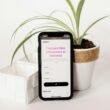 Ein Mobiltelefon liegt neben einem grünen Blumentopf auf einem Holztisch in einem hellen Innenraum, modernes Setting