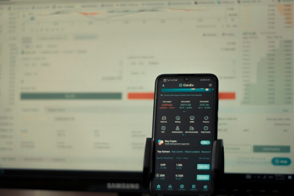 Ein Mobiltelefon steht zentral vor einem großen Computerbildschirm auf einem Schreibtisch, Technik-Alltag im Büro.