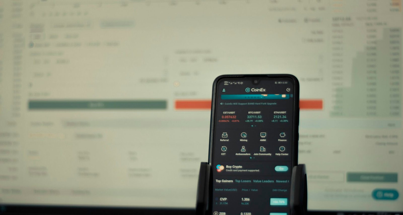 Ein Smartphone liegt vor einem eingeschalteten Computerbildschirm auf dem Schreibtisch, zeigt moderne Arbeitsweise.
