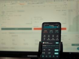 Ein Smartphone liegt vor einem eingeschalteten Computerbildschirm auf dem Schreibtisch, zeigt moderne Arbeitsweise.