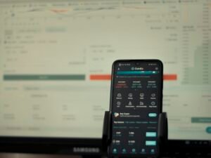 Ein Smartphone liegt vor einem eingeschalteten Computerbildschirm auf dem Schreibtisch, zeigt moderne Arbeitsweise.
