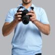 Ein Mann steht mit einer Kamera in der Hand vor neutralem Hintergrund – Symbolbild für Fotografie und Technik.