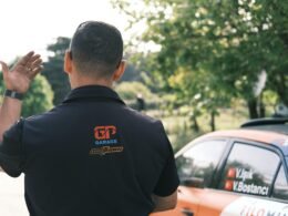 Ein Mann steht neben einem geparkten Auto auf einer Straße in städtischer Umgebung, fotografiert bei Tageslicht.