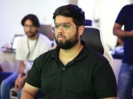 Ein Mann mit Brille sitzt an einem Tisch und spricht vor einer Gruppe von Personen, die aufmerksam zuhören.