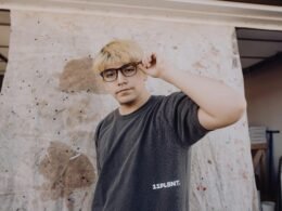 Ein Mann mit Brille steht aufrecht vor einer hellen, neutralen Wand und blickt nach vorne.