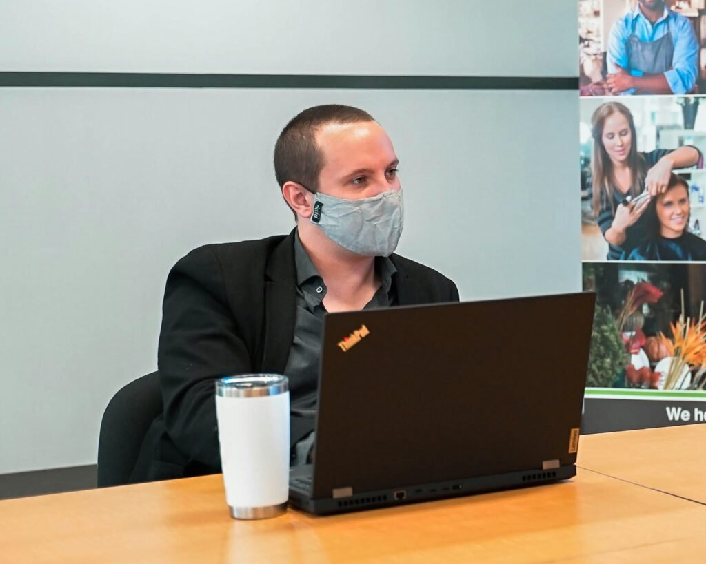 Ein Mann mit Gesichtsmaske sitzt vor einem Laptop an einem Schreibtisch in einem modernen Arbeitsumfeld.