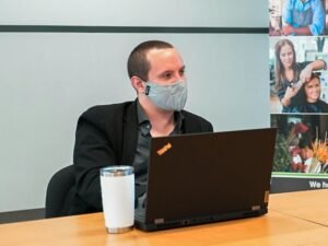 Ein Mann mit Gesichtsmaske sitzt vor einem Laptop an einem Schreibtisch in einem modernen Arbeitsumfeld.