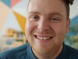 Ein Mann mit roten Haaren blickt lächelnd direkt in die Kamera, neutrale Kulisse, freundlicher Ausdruck