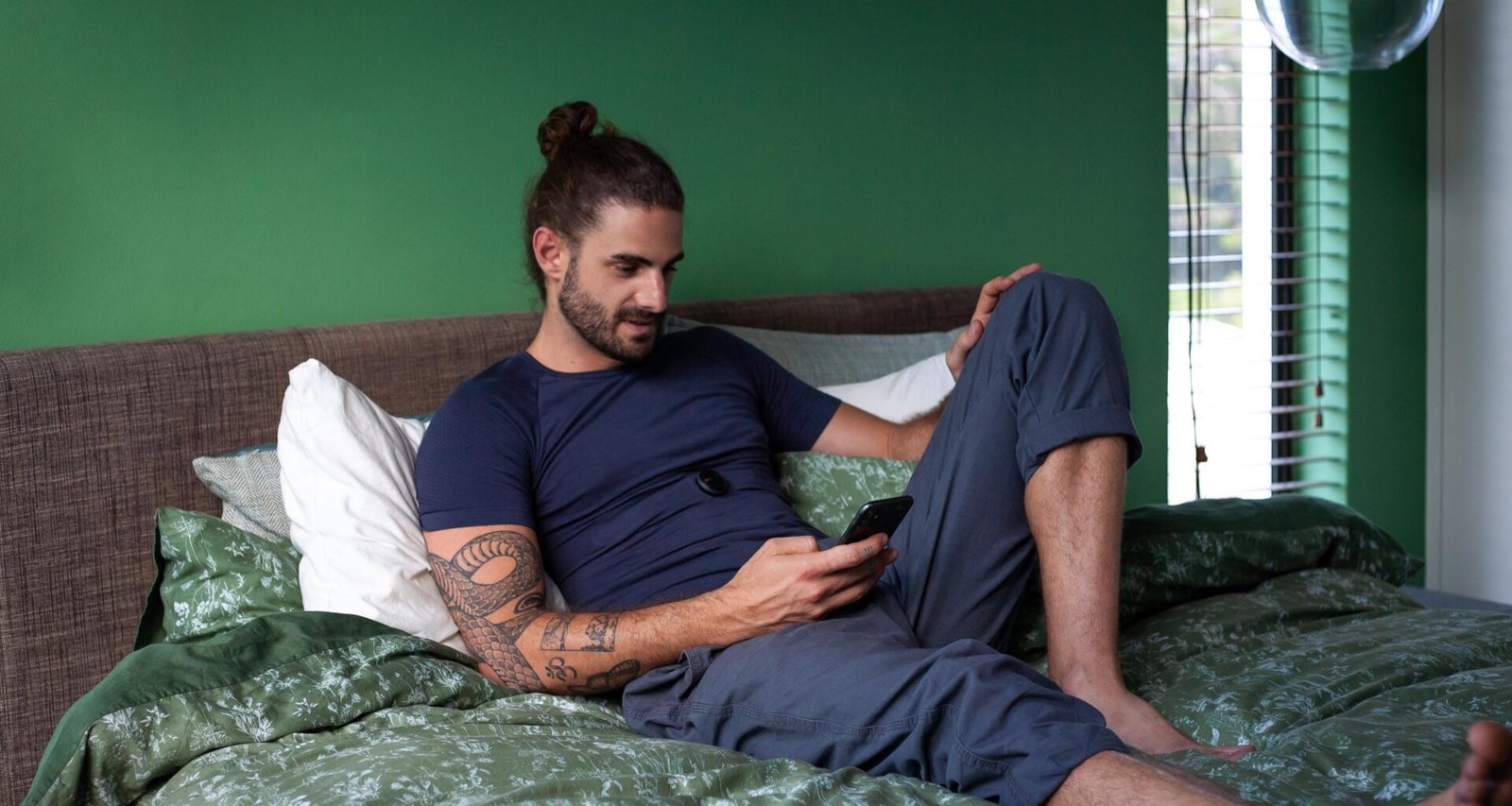 Ein Mann sitzt auf einem Bett und schaut konzentriert auf sein Handy, das Schlafzimmer ist minimalistisch eingerichtet.