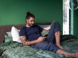 Ein Mann sitzt auf einem Bett und schaut konzentriert auf sein Handy, das Schlafzimmer ist minimalistisch eingerichtet.