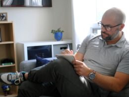 Ein Mann sitzt entspannt auf einer hellgrauen Couch im Wohnzimmer, Konzept für flexibles Arbeiten von zu Hause aus.