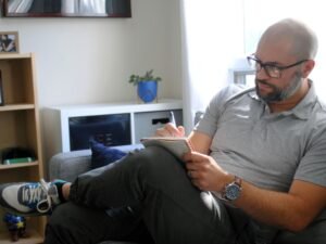 Ein Mann sitzt entspannt auf einer hellgrauen Couch im Wohnzimmer, Konzept für flexibles Arbeiten von zu Hause aus.