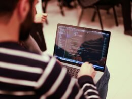 Ein Mann sitzt an einem Tisch vor einem aufgeklappten Laptop und arbeitet konzentriert am Computer