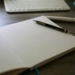 Ein Notizbuch mit einem Stift darauf liegt neben einer weißen Computertastatur auf einem Schreibtisch.
