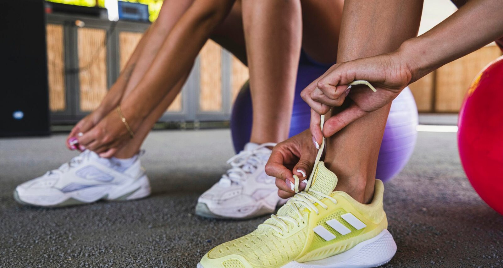Zwei Personen sitzen nebeneinander und binden ihre Schnürsenkel an Sportschuhen auf einem Gehweg