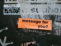 Ein Schild mit der Aufschrift 'Message for you?' ist an einer grauen Wand befestigt und thematisiert Kommunikation.