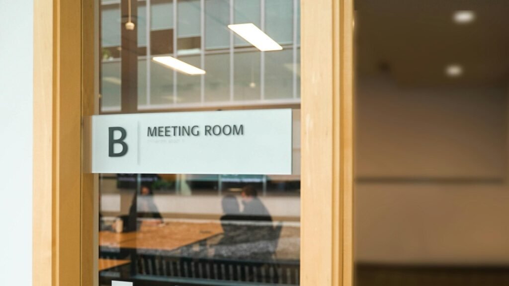Ein Schild mit der Aufschrift Meeting Room hängt an einer Glastür und kennzeichnet einen Besprechungsraum in einem modernen Büro.