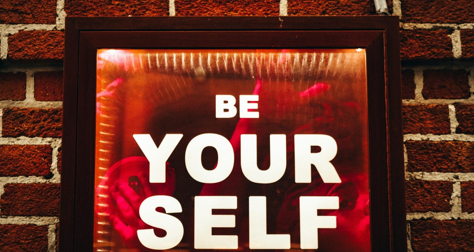 Schild mit der Aufschrift Be Yourself angebracht an einer roten Backsteinwand, aufgenommen von Jon Tyson auf Unsplash