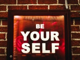 Schild mit der Aufschrift Be Yourself angebracht an einer roten Backsteinwand, aufgenommen von Jon Tyson auf Unsplash