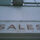 Ein rotes Verkaufsschild mit Aufschrift 'Sales' ist deutlich an der Außenwand eines Gebäudes angebracht.