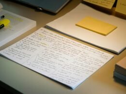 Ein Schreibtisch mit verstreuten Papieren, Kugelschreibern und einem gelb-grünen Textmarker zur Arbeitsorganisation