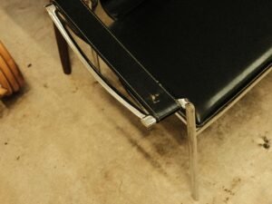 Ein schwarzer Lederstuhl steht direkt neben einem Holzstuhl auf einem schlichten Boden, beide sind leer und gut beleuchtet.